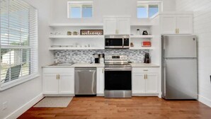 A modern kitchen with white cabinetry, a stainless steel dishwasher, a built-in oven, a microwave, and a refrigerator. There are two windows with blinds, a 'welcome' sign on the shelves, and a tiled backsplash. - Romantic Bungalow "Carmen" on 30 Acres (Dripping Springs)