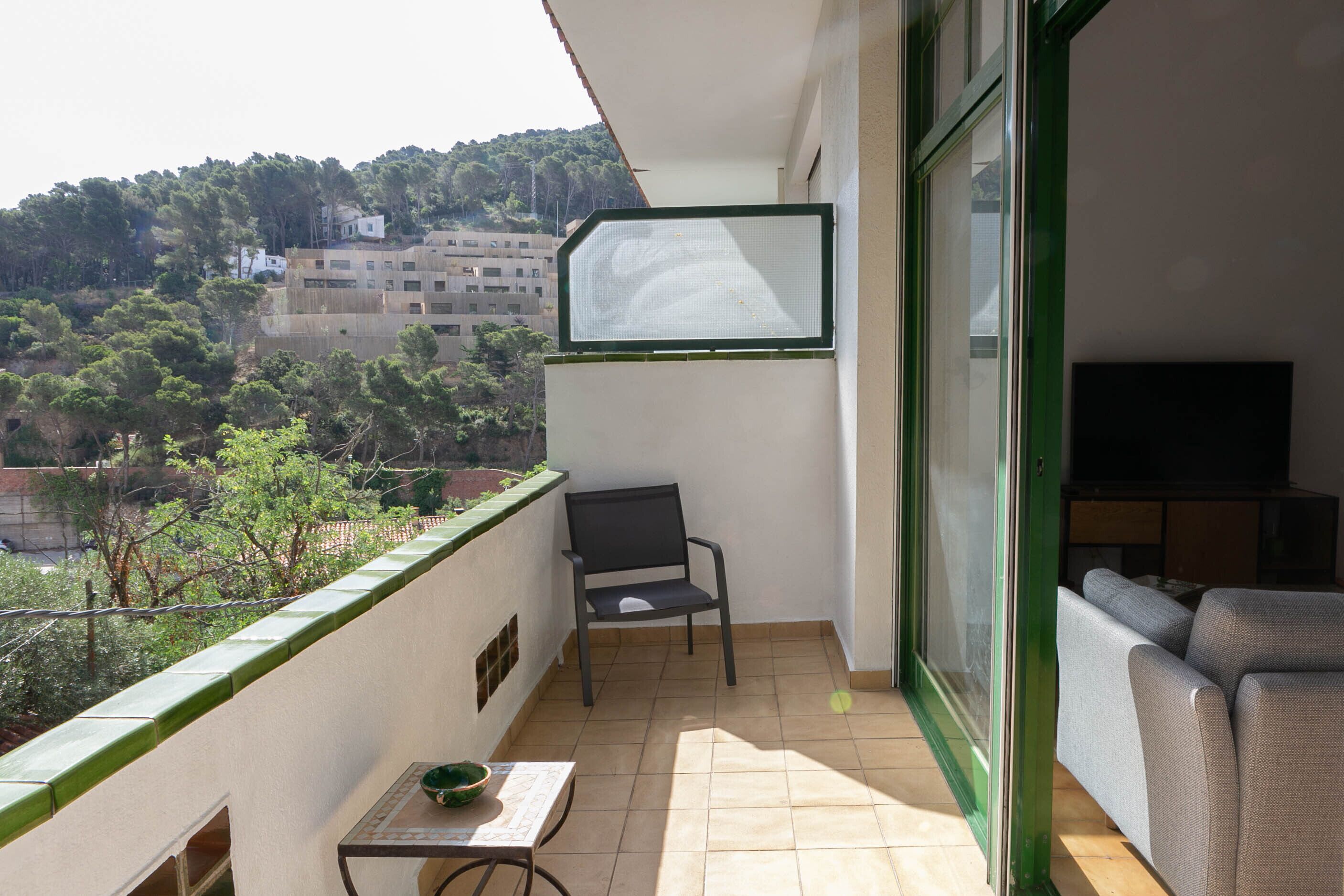 A balcony with a chair, a small table, and a sofa. There is a TV mounted on the wall. The balcony overlooks a hillside with buildings and trees.
