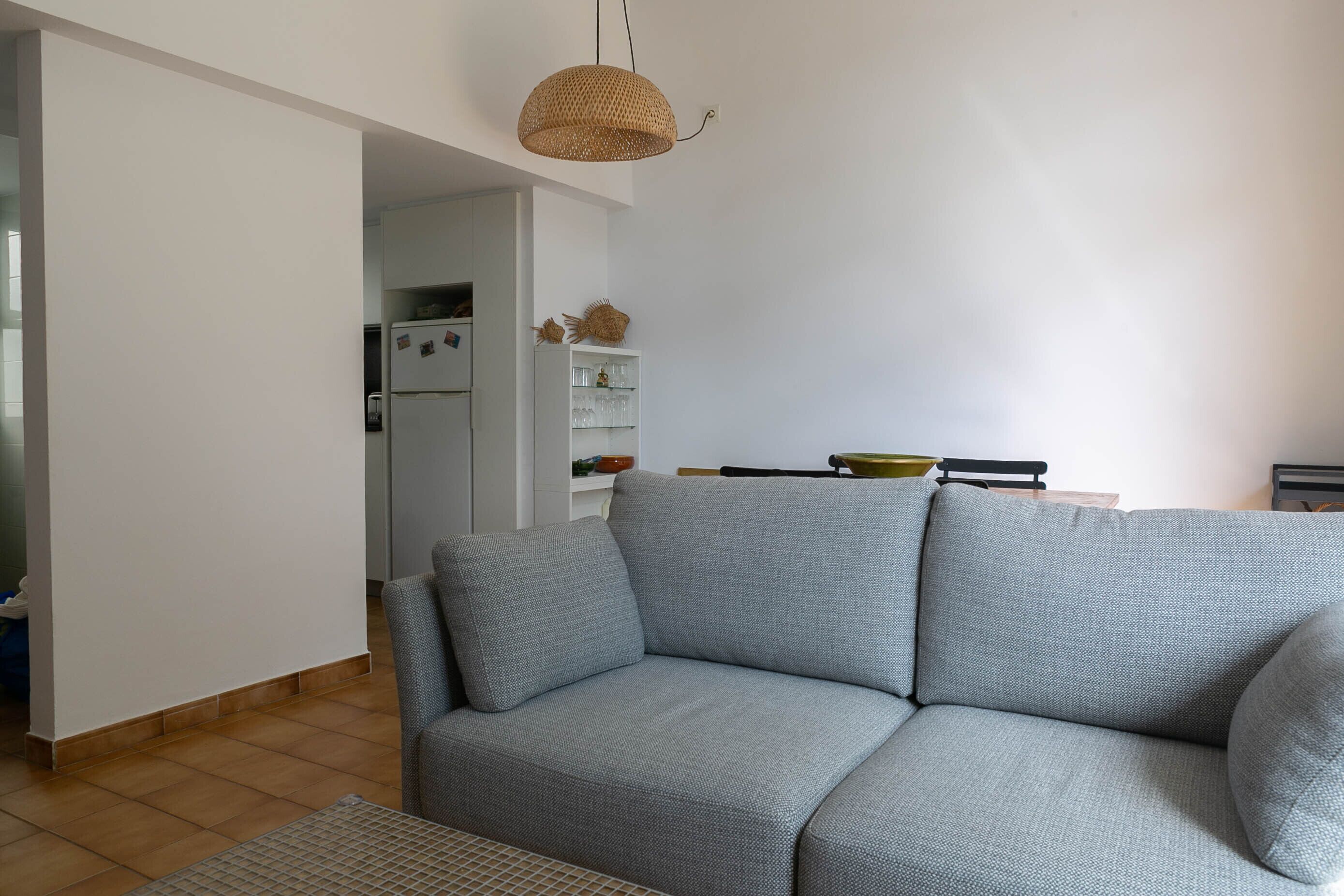 A living room with a grey sofa, a woven pendant light, and a kitchen area in the background.