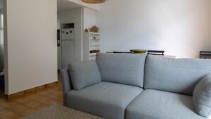 Un salon avec un canapé gris, un luminaire suspendu en fibres tressées et, au fond, un coin cuisine.