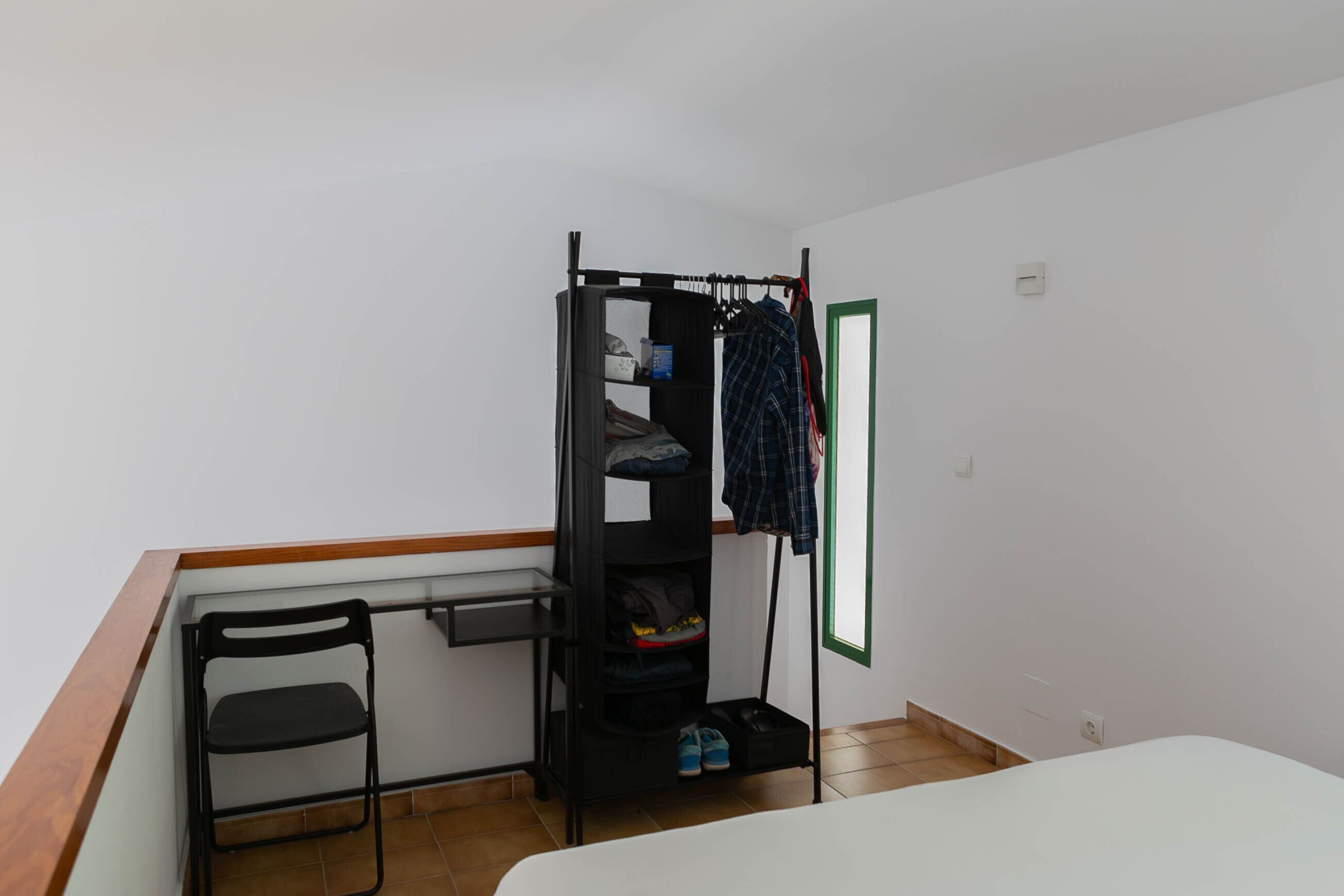 A small room with a bunk bed, a chair, and a shelf with clothes and shoes.
