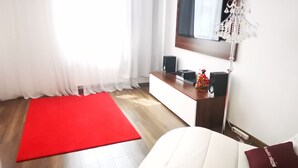 A modern hotel room with a wooden floor, a red carpet, a white sofa, a TV mounted on the wall, and a small table with speakers and a decorative item. - Apartament Ogarna II (Gdansk)