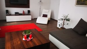 A modern living room with a wooden coffee table, red candles, a white sofa, a white armchair, a red rug, a potted plant, and a flat-screen TV mounted on the wall. - Apartament Ogarna II (Gdansk)