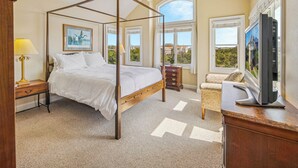 A four-poster bed with a wooden frame, a television on a wooden stand, and a view of greenery through the windows. - Oceanside Dog Friendly Home w/ Hot Tub, Game Room, and Wet Bar. Island Stylin'! (Frisco)