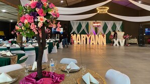 Una sala da banchetto con tavoli rotondi apparecchiati per un evento formale, con un'ampia composizione floreale al centro, tende bianche drappeggiate e un palco con un grande cartello illuminato.