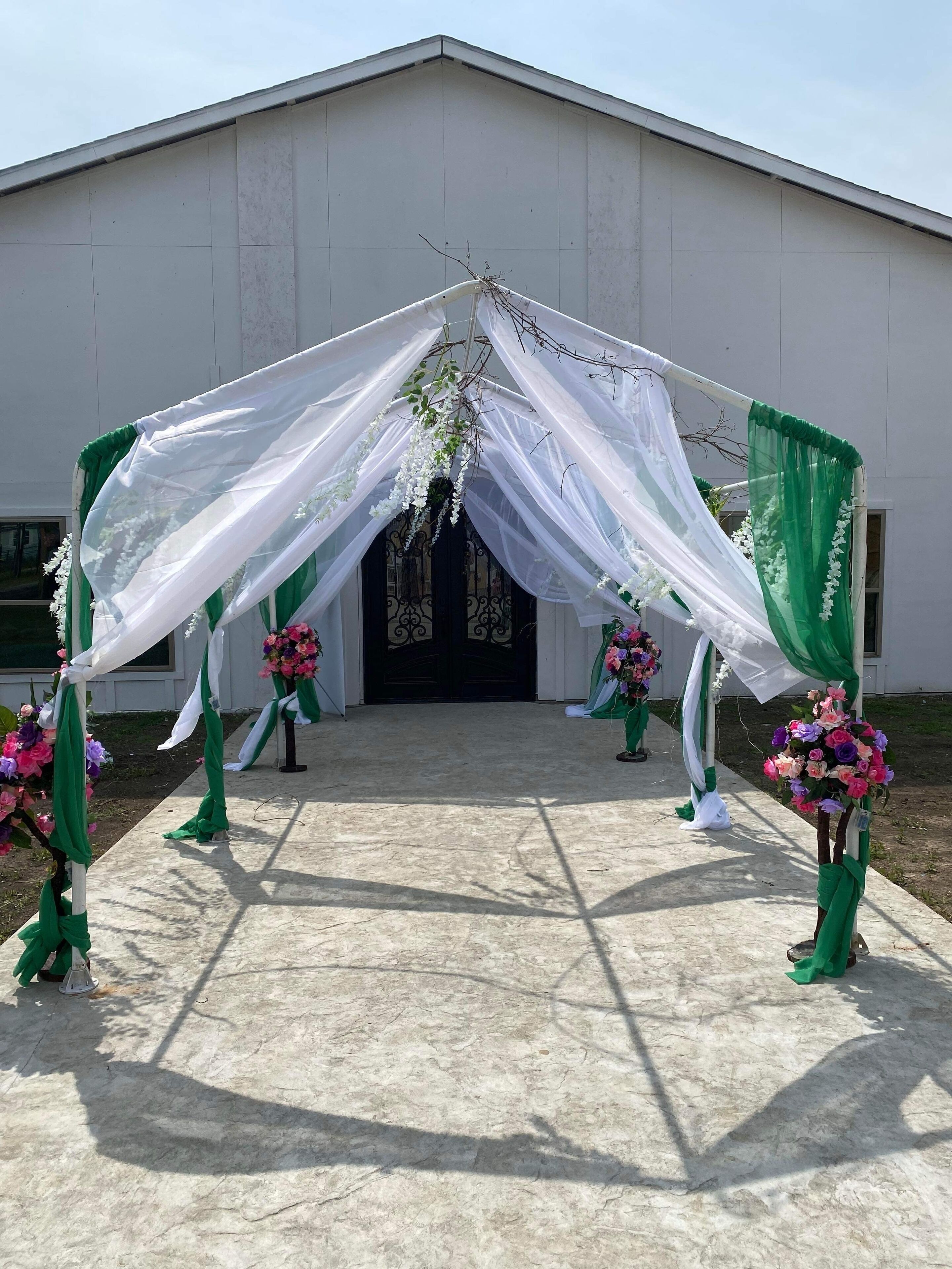 An outdoor wedding arch with white and green drapes, floral arrangements, and a building in the background.