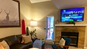 Ein Wohnzimmer mit Kamin, einem an der Wand befestigten Fernseher, einer Couch, zwei Sesseln, einem Couchtisch und einem großen Bild von Hirschen.