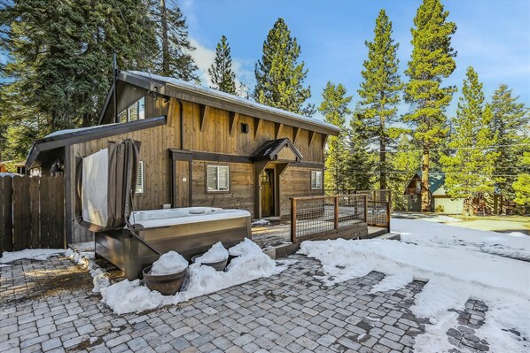 A wooden cabin with a hot tub, surrounded by snow and trees.