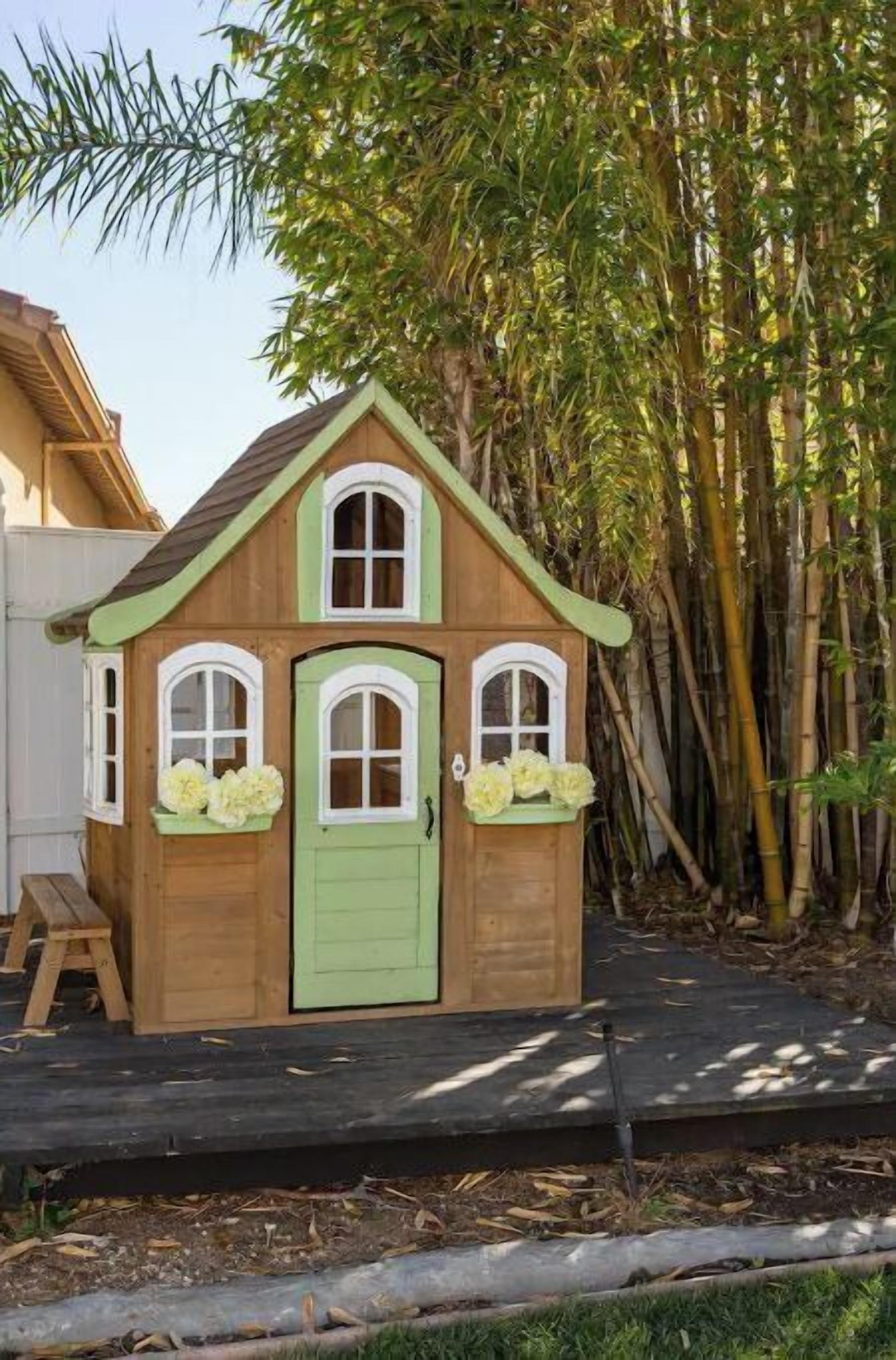 A small wooden playhouse with a green door and windows, surrounded by bamboo plants.