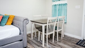 Una habitación pequeña y ordenada con un sofá gris, una mesa de comedor blanca con cuatro sillas y una ventana con cortinas color turquesa.