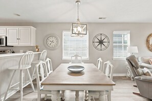 A dining area with a wooden table, white chairs, a wall clock, and a decorative wall piece.