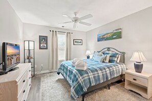 A bedroom with a bed, a TV, a ceiling fan, a window with curtains, and a painting on the wall.