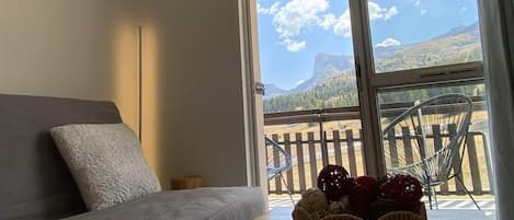 A room with a sofa, a window with a view of mountains, and a basket of fruit on a table.