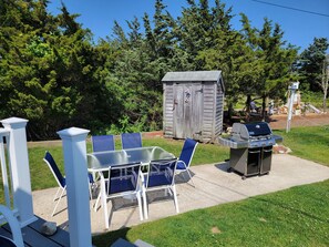 An outdoor patio with a glass table and chairs, a barbecue grill, and a wooden shed.