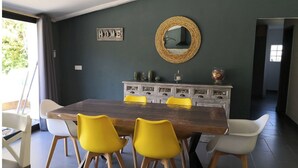 Uma área de refeições com uma mesa de madeira e cadeiras amarelas, um armário cinzento com objetos decorativos e um espelho na parede.