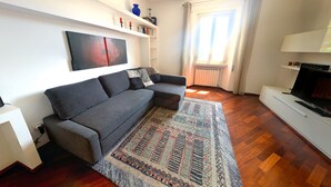 A modern living room with a grey sofa, a patterned rug, wooden flooring, a white entertainment unit, and a window with curtains. - New Bright and Quiet in the Heart of Trastevere (Roma)