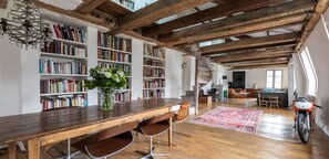 A spacious room with exposed wooden beams, a long wooden table with chairs, bookshelves filled with books, a decorative vase with flowers, and a motorcycle in the corner. - ON THE BANKS OF SEINE-AN EXPLICIT 17TH CENT.TOWNHOUSE LE MARAIS VILLAGE ST PAUL (Paris)