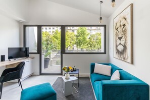 A modern living room with a teal sofa, a flat-screen TV, a desk, and a large window offering a view of buildings and trees.
