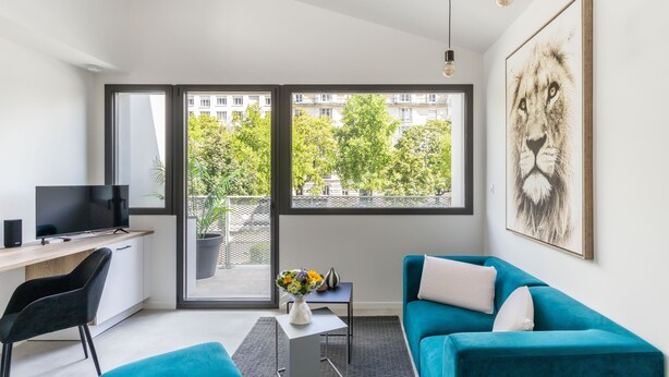 A modern living room with a teal sofa, a flat-screen TV, a desk, and a large window offering a view of buildings and trees. - Moines - Appartement moderne et confortable (Rennes)