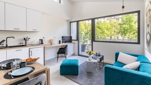 A modern kitchen with white cabinets, a dining table with chairs, a teal sofa, and a large window with a view of trees. - Moines - Appartement moderne et confortable (Rennes)