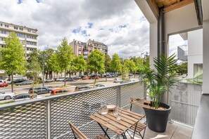 Een balkon met een houten tafel, een bloempot en uitzicht op een straat met auto's en gebouwen.
