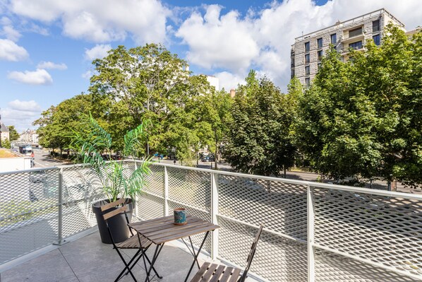 Ein Balkon mit Holztisch, einem Blumentopf und einem Metallgeländer, mit Blick auf eine Straße mit Bäumen und Gebäuden.
