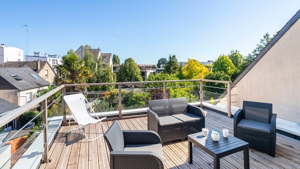 A rooftop terrace with wicker furniture, a glass balustrade, and a view of surrounding buildings and trees. - Teranga - Studio avec grande terrasse dans Rennes (Rennes)