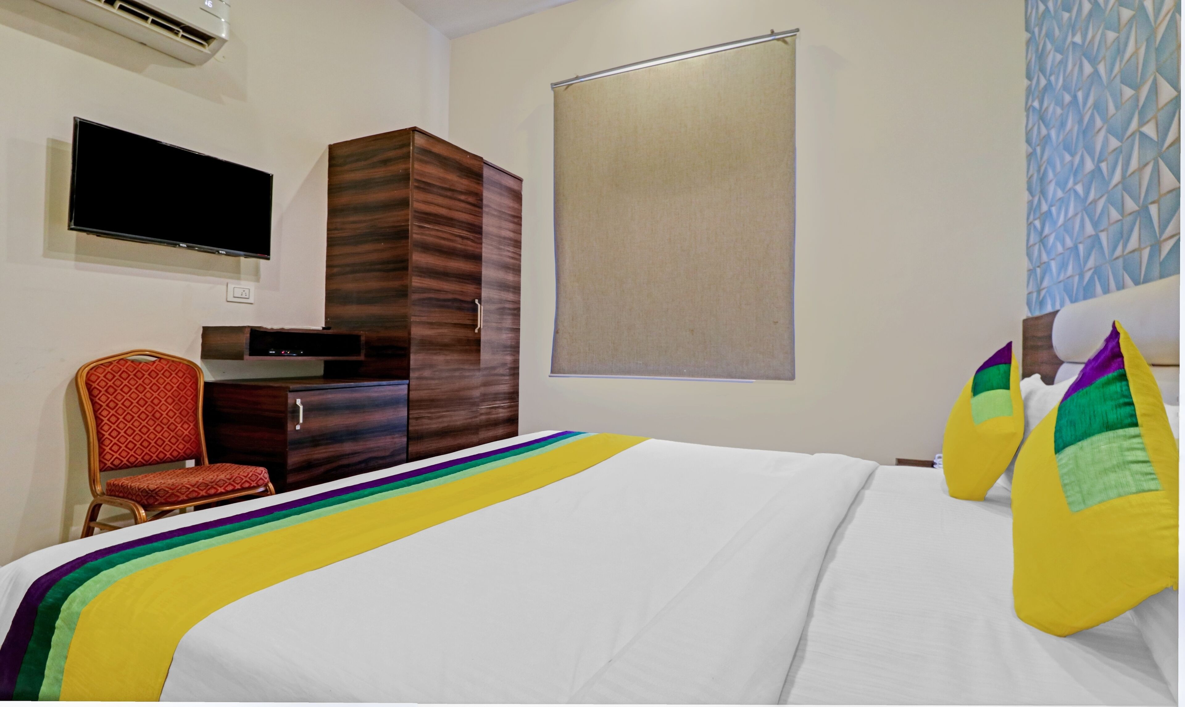 A hotel room with a bed, a TV mounted on the wall, a wardrobe, and a window with a blind.
