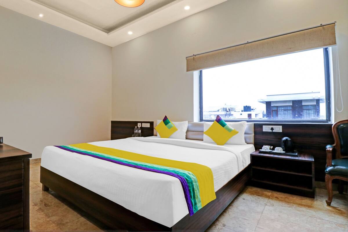 A hotel room with a large bed, a bedside table, a nightstand, a window with a view of buildings, and a ceiling light.