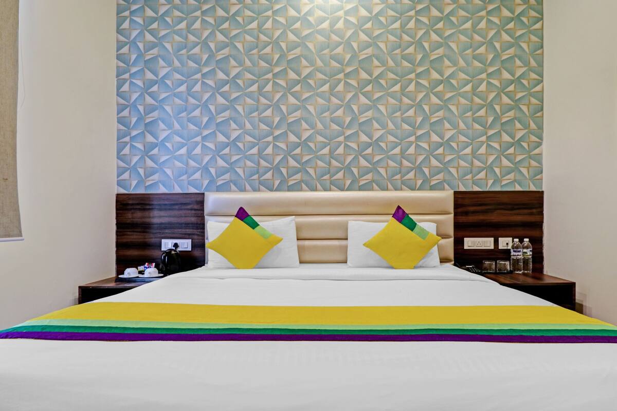 A hotel room with a geometric patterned wall, a bed with a rainbow-striped bedspread, and two pillows with a geometric design.