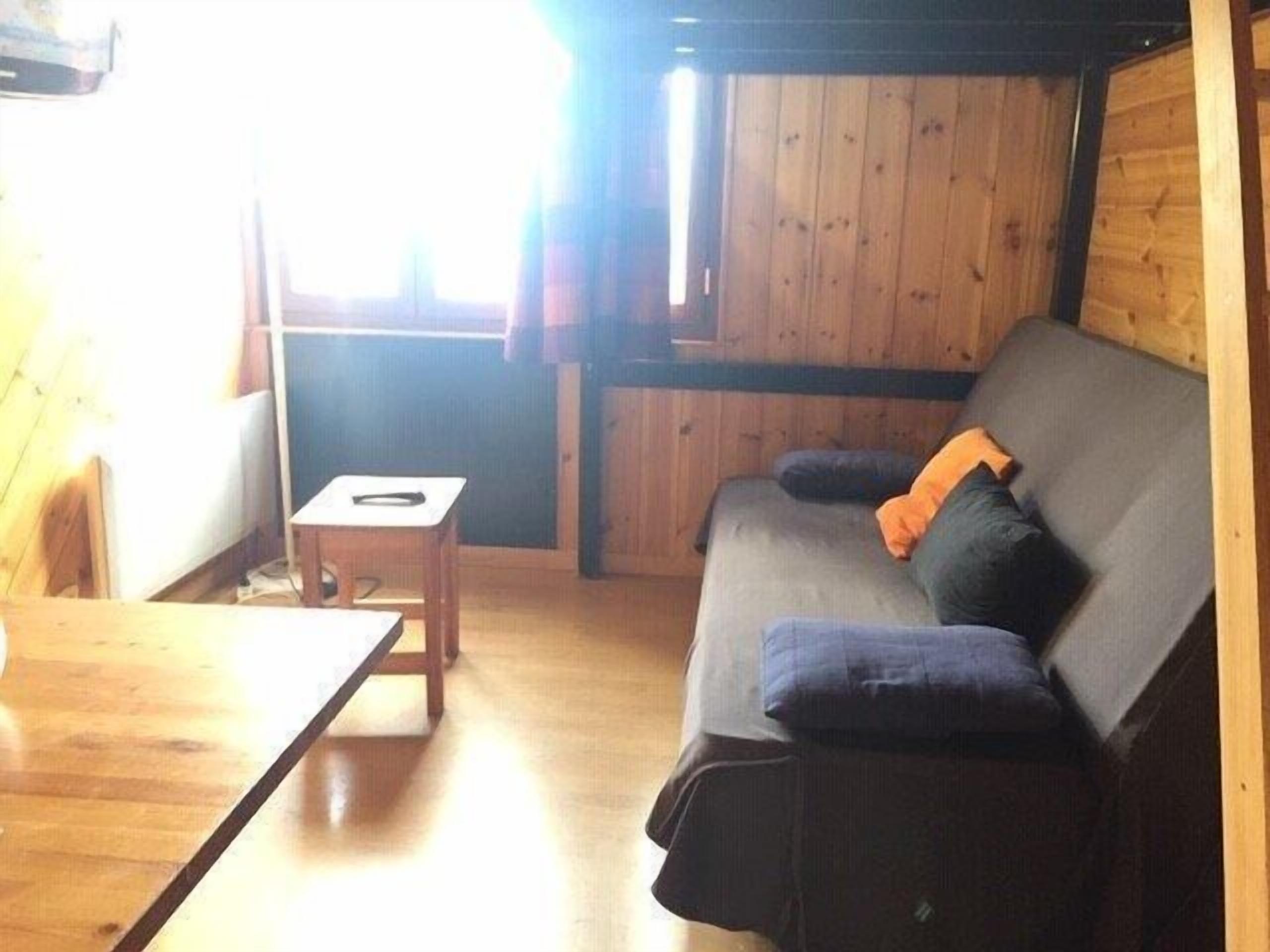 Una habitación acogedora con suelo de madera, un sofá gris con cojines y una pequeña mesa de madera.