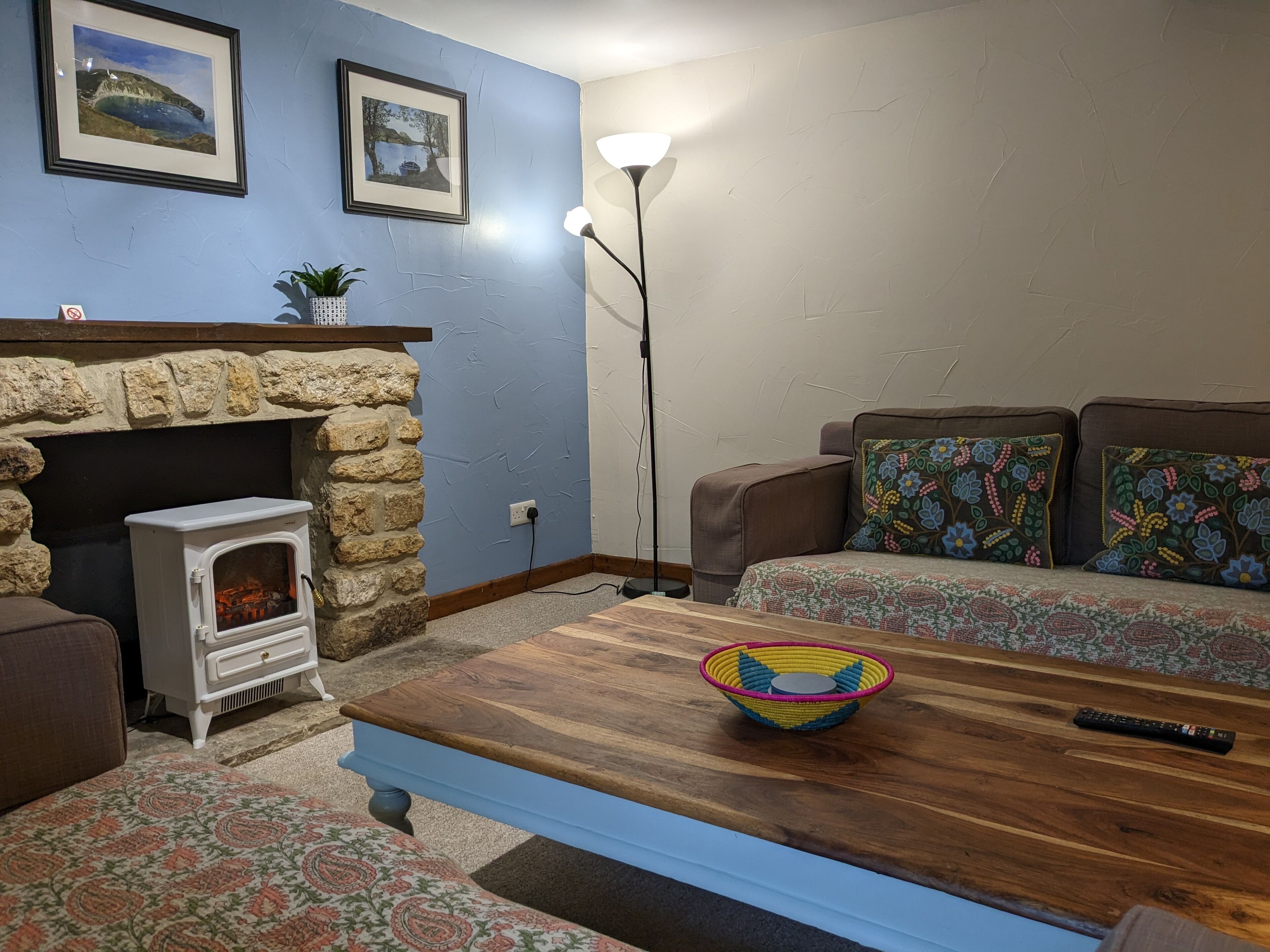 A living room with a stone fireplace, a sofa with patterned cushions, a wooden coffee table, and two framed pictures on the wall.