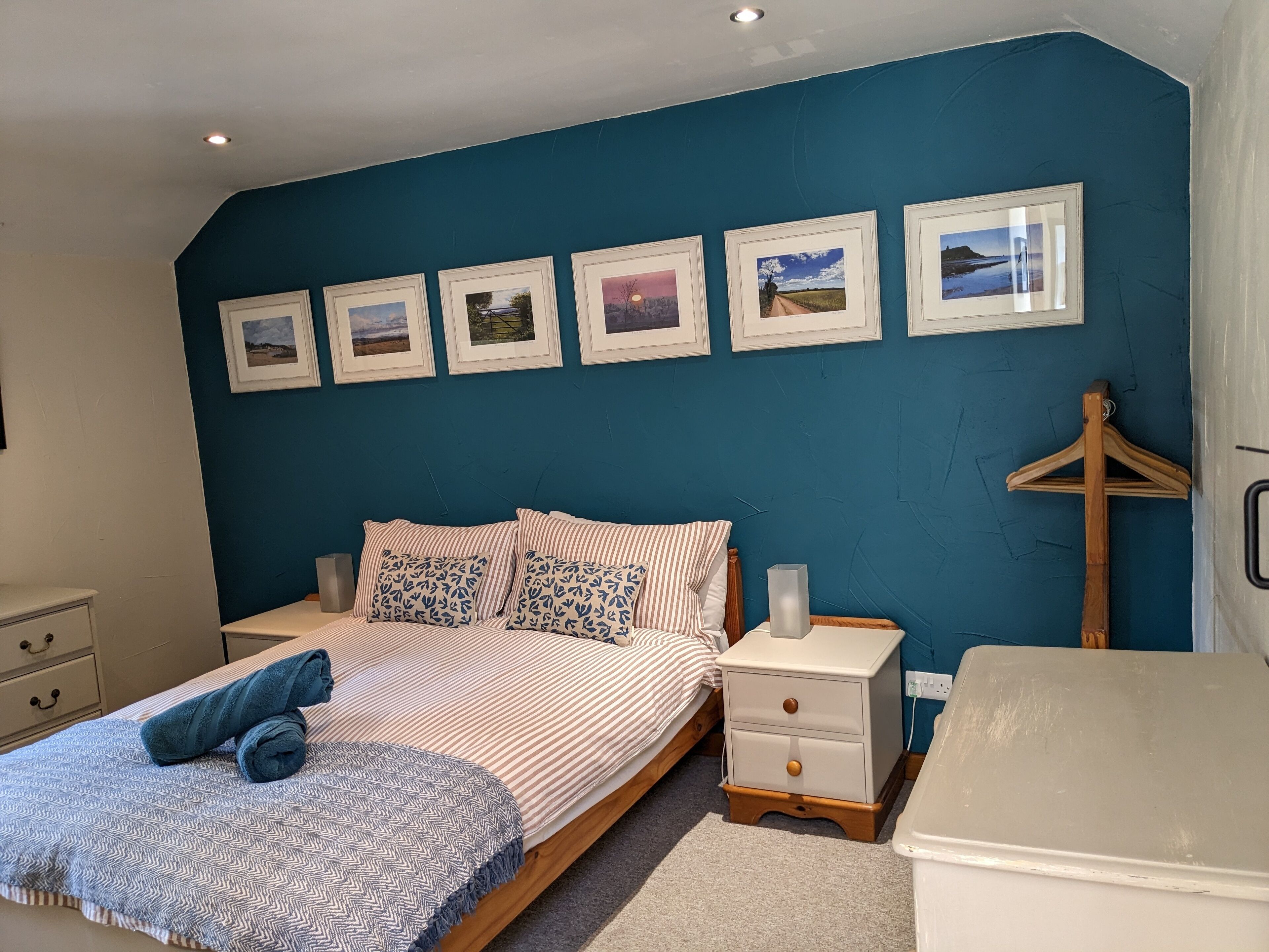 Quarto com cama, mesas de cabeceira, cômoda e parede com quadros.