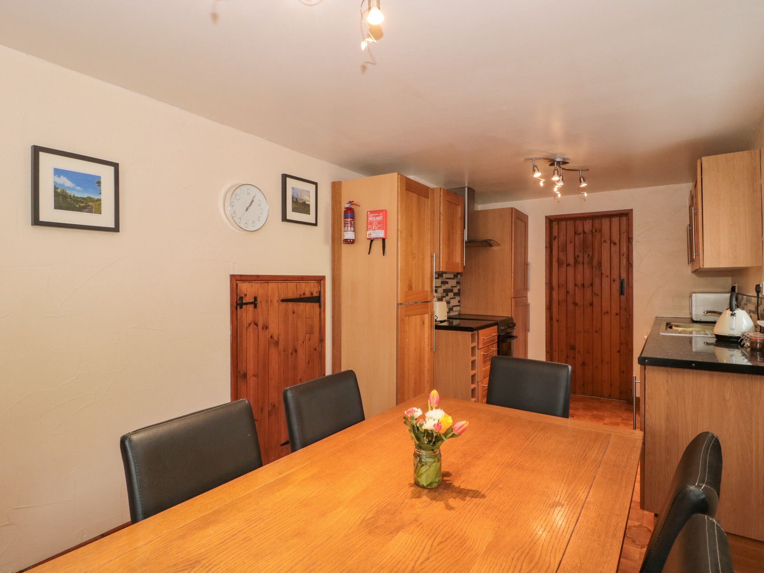 A dining area with a wooden table, chairs, and a vase of flowers. There are wooden cabinets and a kitchen in the background.