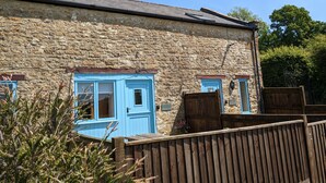 Un bâtiment en pierre avec des portes et des fenêtres bleues, une clôture en bois et un ciel d’un bleu limpide.