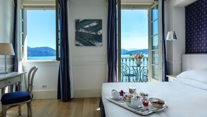 Ein Hotelzimmer mit Bett, Schreibtisch, Stuhl, einem Balkon mit Blick auf Meer und Berge sowie einem Tablett mit Frühstücksartikeln.