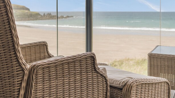 Un fauteuil de relaxation en osier offrant une vue sur la plage et l’océan, visible à travers une fenêtre en verre.