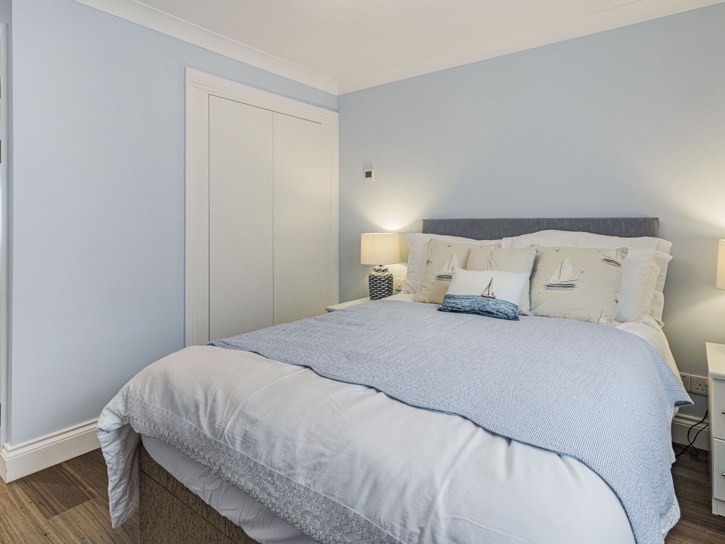 A neatly made bed with a light blue and white duvet, a grey headboard, and multiple pillows with nautical designs. There is a bedside table with a lamp and a decorative basket.
