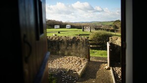 A view from inside a building looking out to a rural landscape with two white caravans parked in a field, a stone wall, and a grassy area with scattered stones. - The Sty (Bridport)