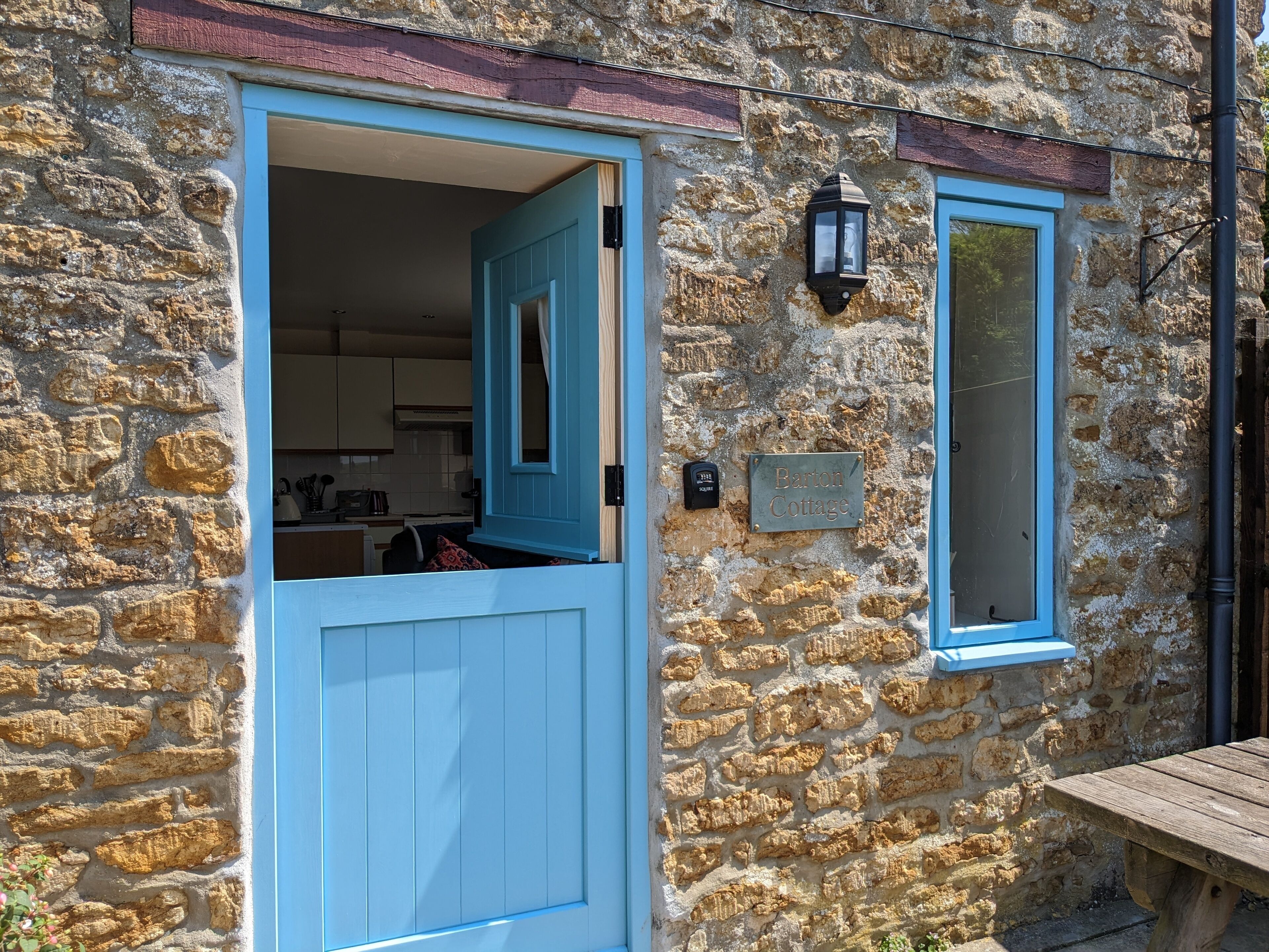 A stone cottage with a blue door and window, a small outdoor table, and a sign reading "Barton Cottage."
