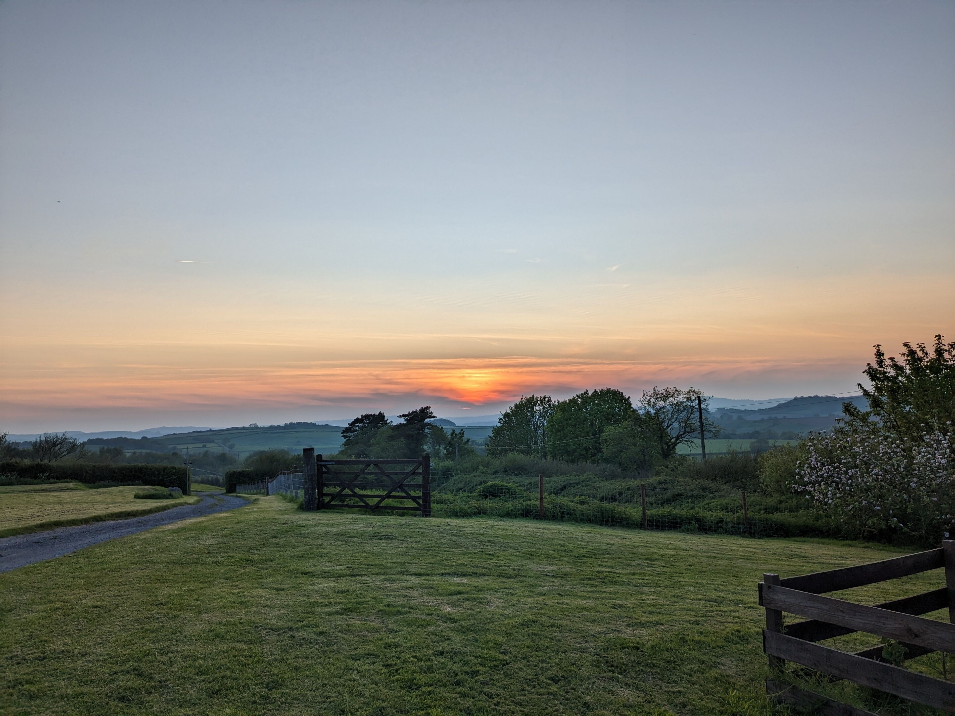 Paisaje rural con una verja de madera, un camino y un atardecer a lo lejos.