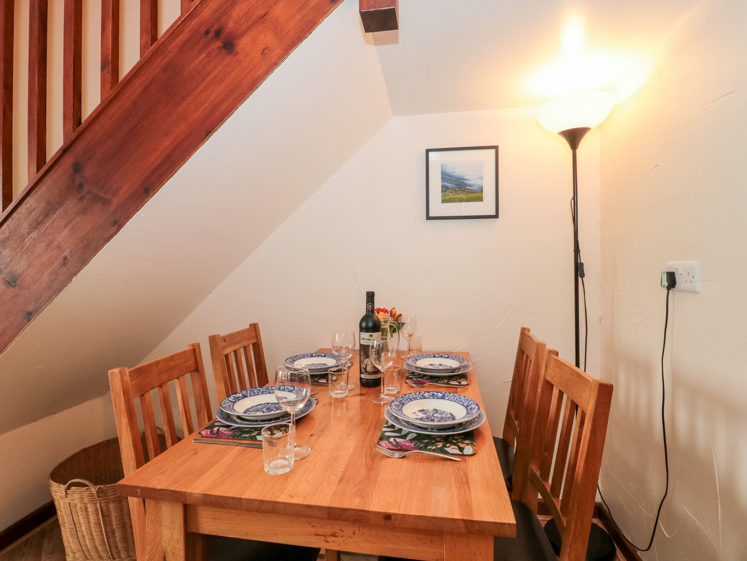 A dining table set with plates, glasses, and a bottle, in a room with a wooden staircase and a wall-mounted lamp.