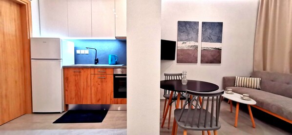 Een kleine keuken met een koelkast, spoelbak en waterkoker, plus een zithoek met een bank, salontafel en kunstwerk.