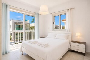 Una habitación de hotel moderna con una cama grande, una mesita de noche con lámpara, una ventana con cortinas y un balcón con vistas a edificios y palmeras.