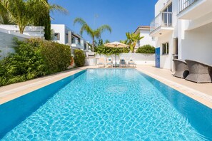Una piscina con sillones de descanso, una pérgola y un edificio blanco con balcones.