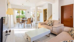 A spacious living area with a sofa, coffee table, and dining table. There is a TV, lamp, and a view of the outside. - Villa Marina - Three Bedroom Villa, Sleeps 6 (Pernera)