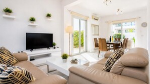 A modern living room with a flat-screen TV, beige sofas, a coffee table, and a dining area with chairs and a table. - Villa Marina - Three Bedroom Villa, Sleeps 6 (Pernera)