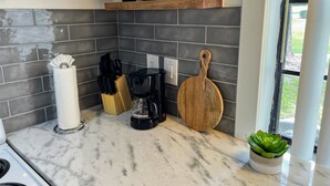 A kitchen with grey tiled backsplash, a wooden shelf with black bowls, a coffee maker, a cutting board, and a potted plant.