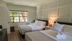 A hotel room with two beds, a window with a view of palm trees, a bedside lamp, and a small plant on the windowsill.