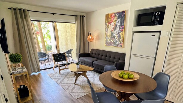 A modern living room with a sofa, a dining table with chairs, a kitchen area with a microwave, and a large window with curtains.